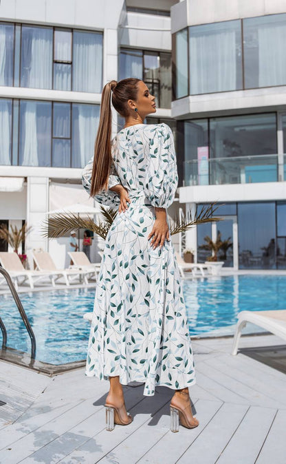 Woman in a white dress with green patterns standing by a poolside.