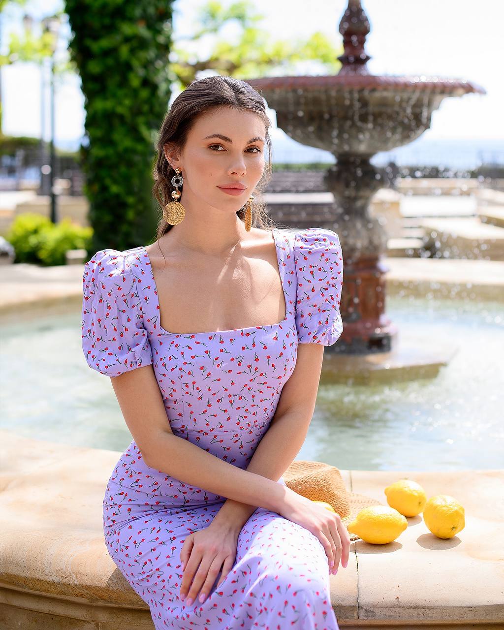 Woman in a light purple dress sitting by a fountain with lemons on a stone ledge.