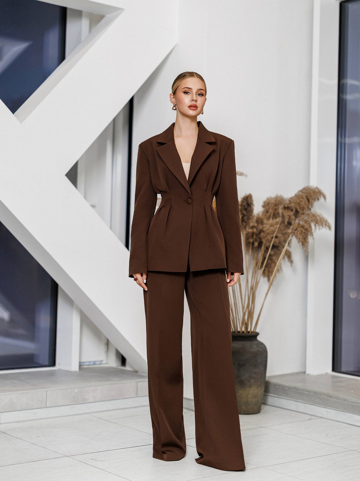 Woman in a brown suit standing in a modern indoor setting with white walls and geometric design.