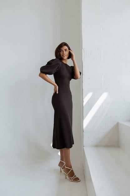 Woman in a black dress standing against a white wall with sunlight casting shadows.