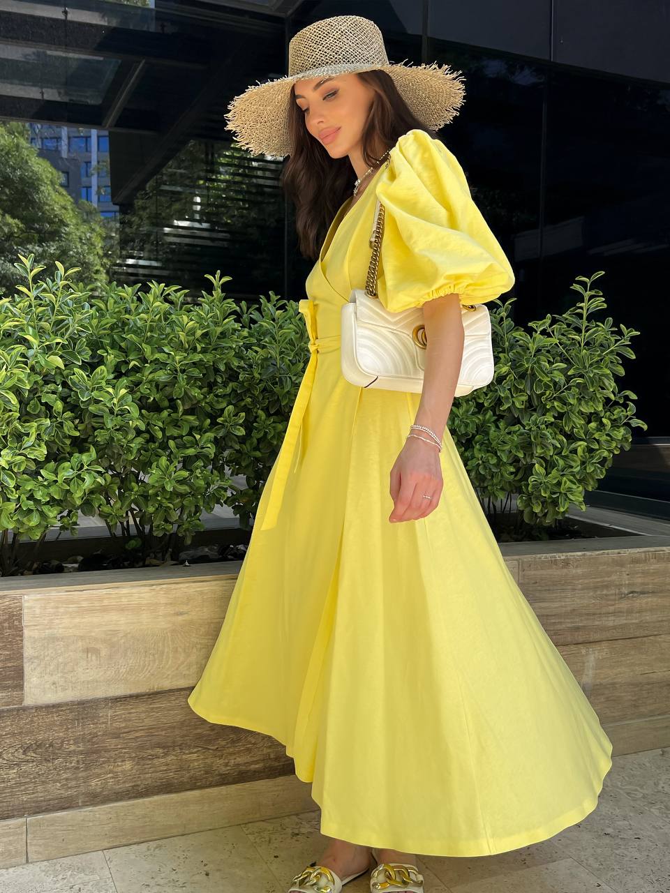 Woman in a yellow dress and straw hat standing outdoors with greenery in the background