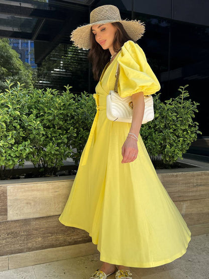 Woman in a yellow dress and straw hat standing outdoors with greenery in the background