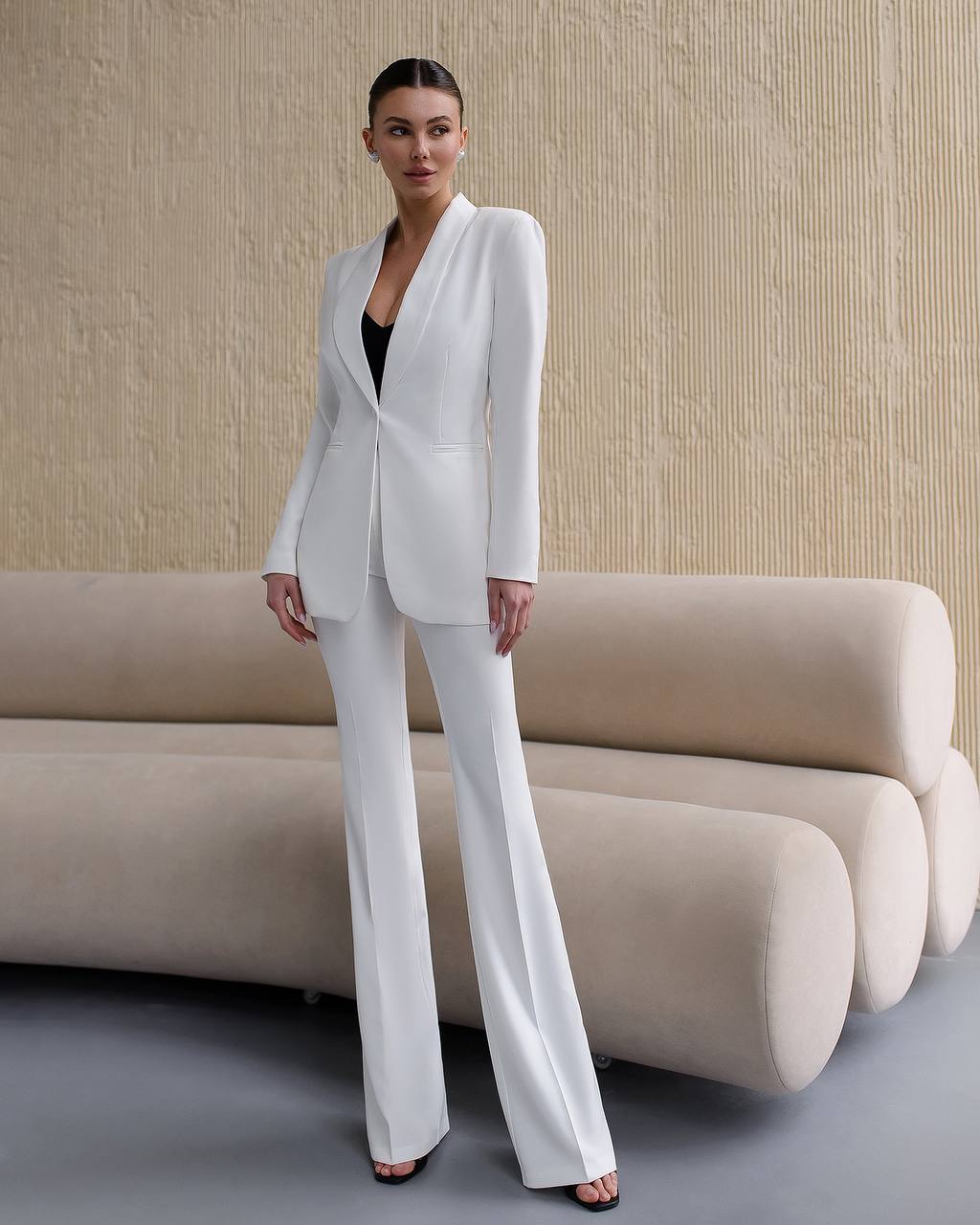 Woman in a white suit standing in a modern interior setting with a beige sofa.