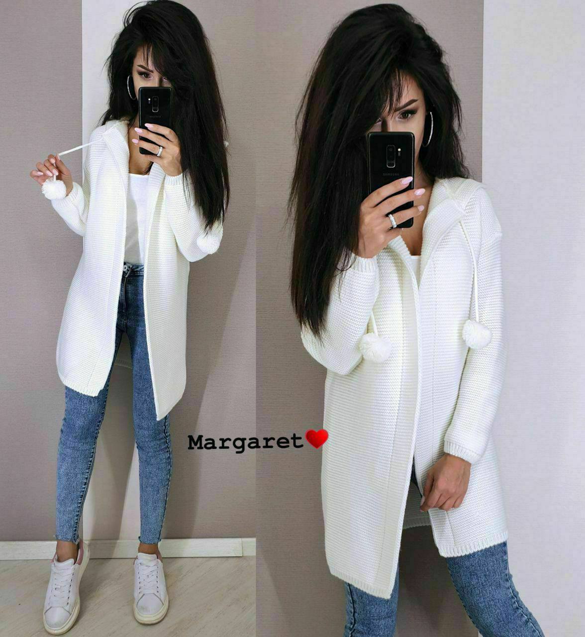 Woman wearing a white cardigan and blue jeans, taking a mirror selfie.