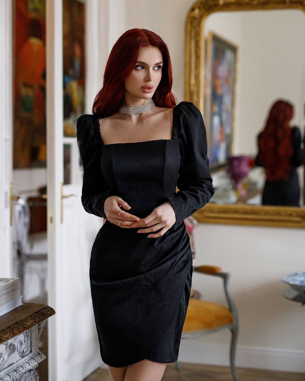 Woman in a black dress standing in an elegant room with mirrors and artwork.