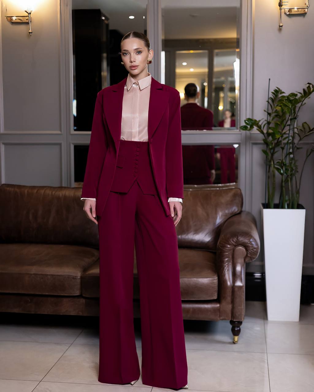 Woman in a burgundy suit standing in an elegant room with a brown leather sofa and decorative elements.