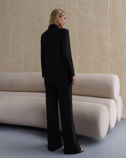 Woman in a black suit standing in front of a beige sofa against a textured wall.