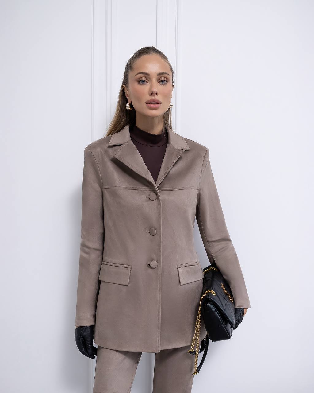 Woman wearing a brown coat and pants with a black handbag against a white background