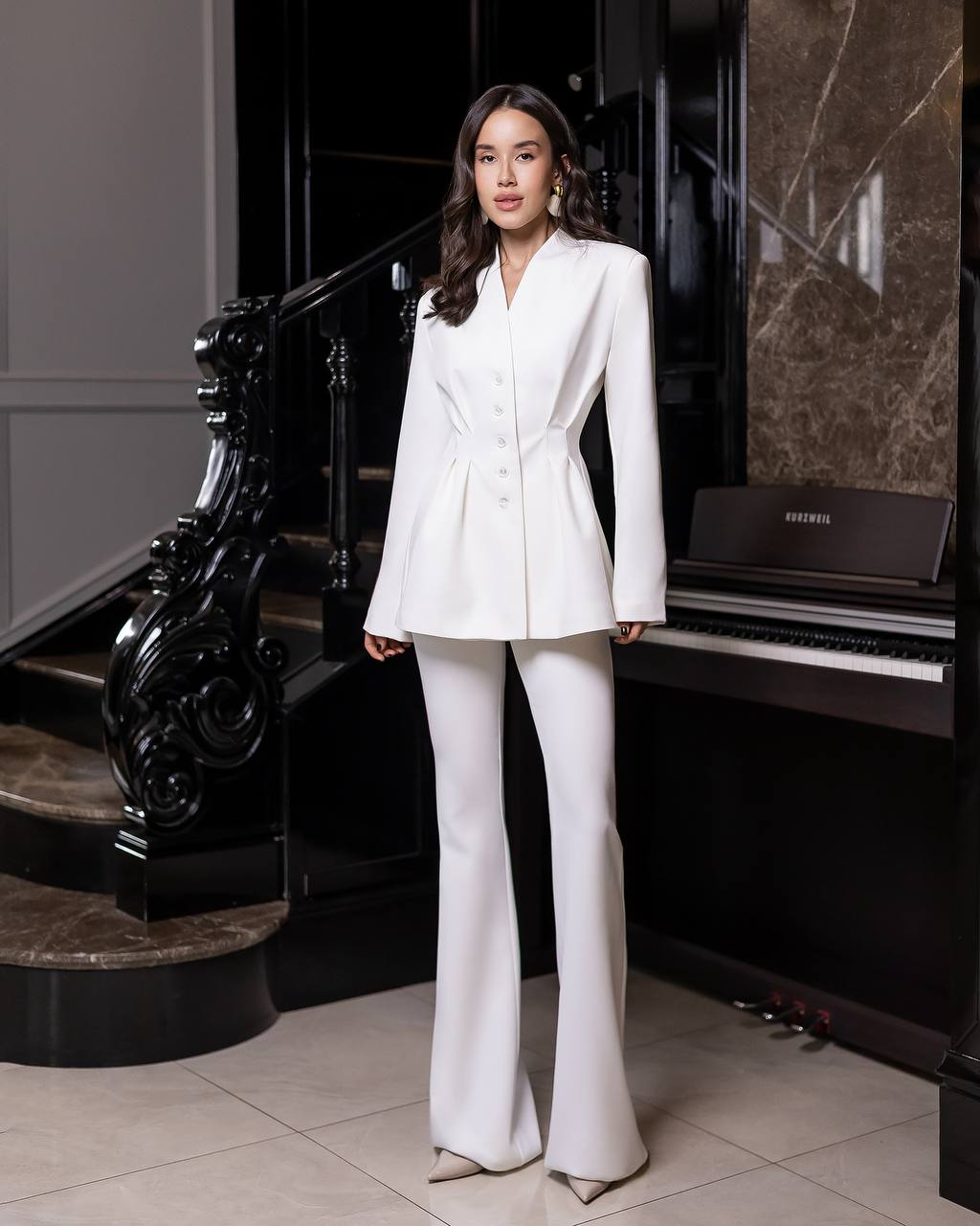 Woman in a white suit standing in a stylish interior setting with a piano.