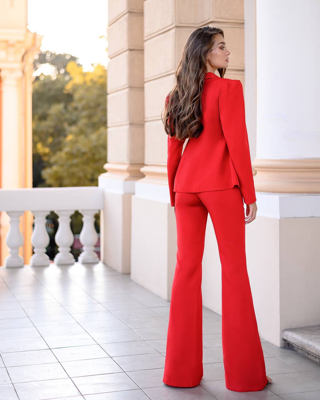 Woman in a red suit standing on a balcony with classical architecture.