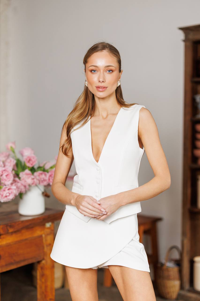 Woman wearing a white dress standing in a room with flowers and furniture.