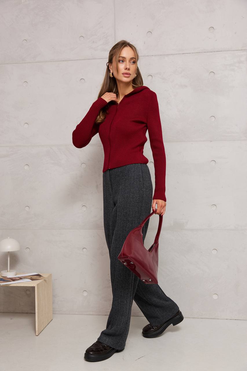 Woman in a red sweater and gray pants holding a red handbag against a light gray wall.
