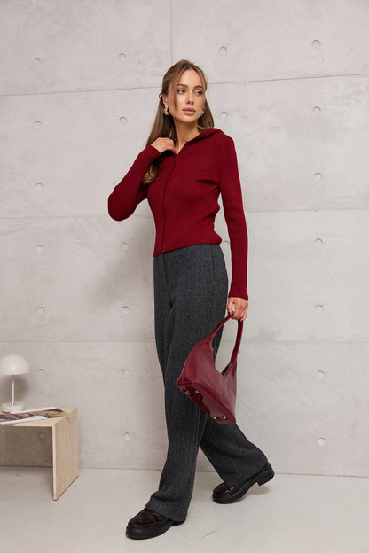 Woman in a red sweater and gray pants holding a red handbag against a light gray wall.