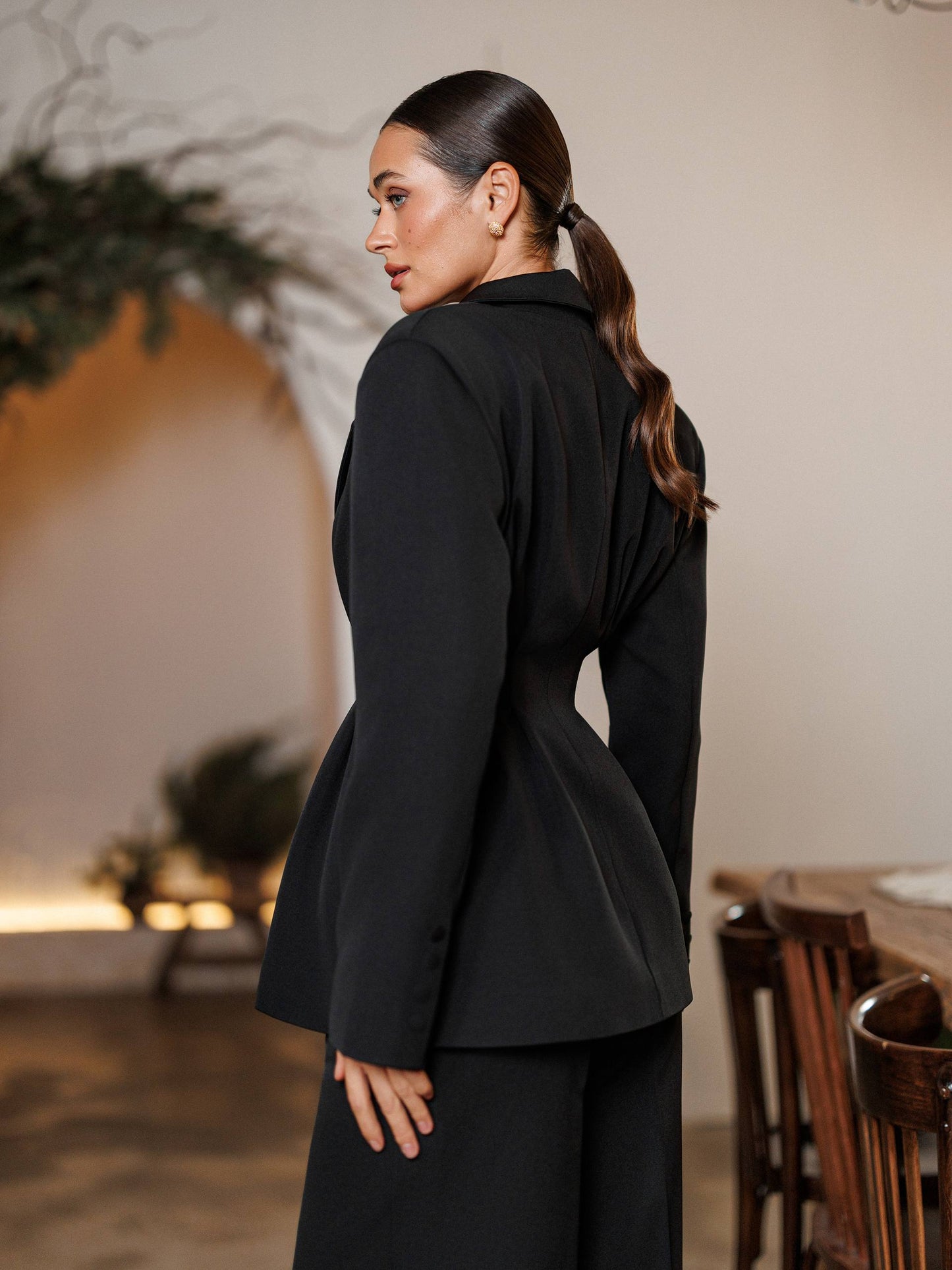 Woman wearing a black blazer in an indoor setting with plants and wooden furniture.