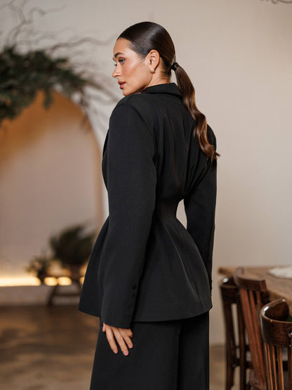 Woman wearing a black blazer in an indoor setting with plants and wooden furniture.