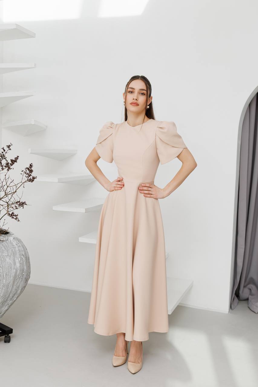 Woman wearing a beige dress in a minimalistic room with white walls and a gray vase.