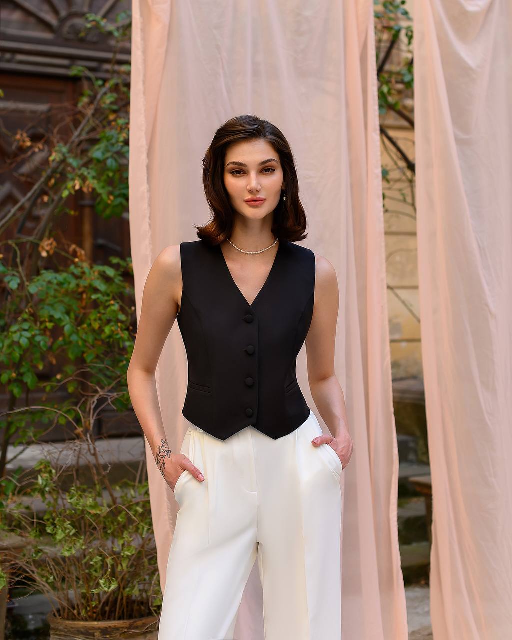 Woman wearing a black sleeveless top and white pants standing in front of a pink curtain.