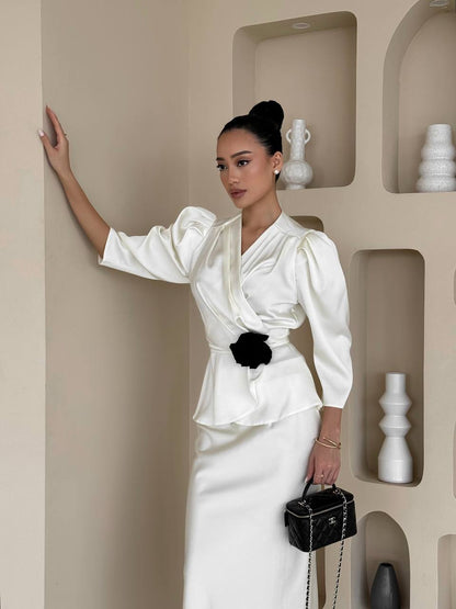 Woman in a white outfit with a black belt standing against a beige wall with decorative shelves.