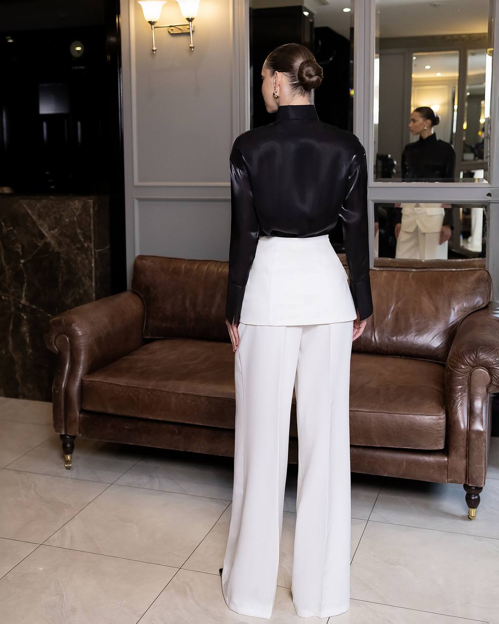 Woman in black top and white pants standing in a room with a brown leather couch.
