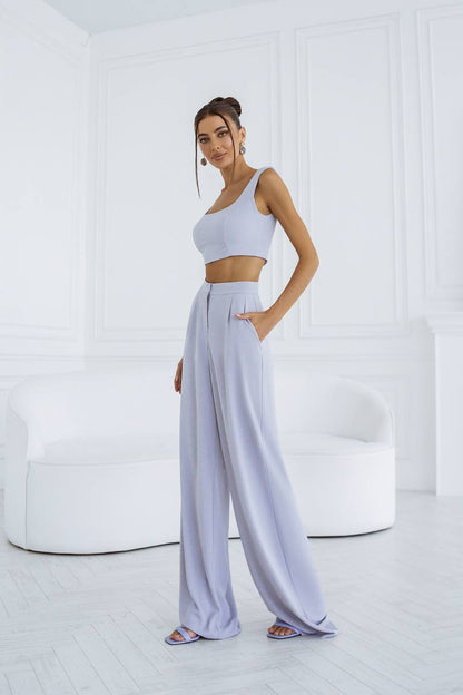 Woman wearing a light gray one-shoulder top and wide-leg pants in a minimalistic room.