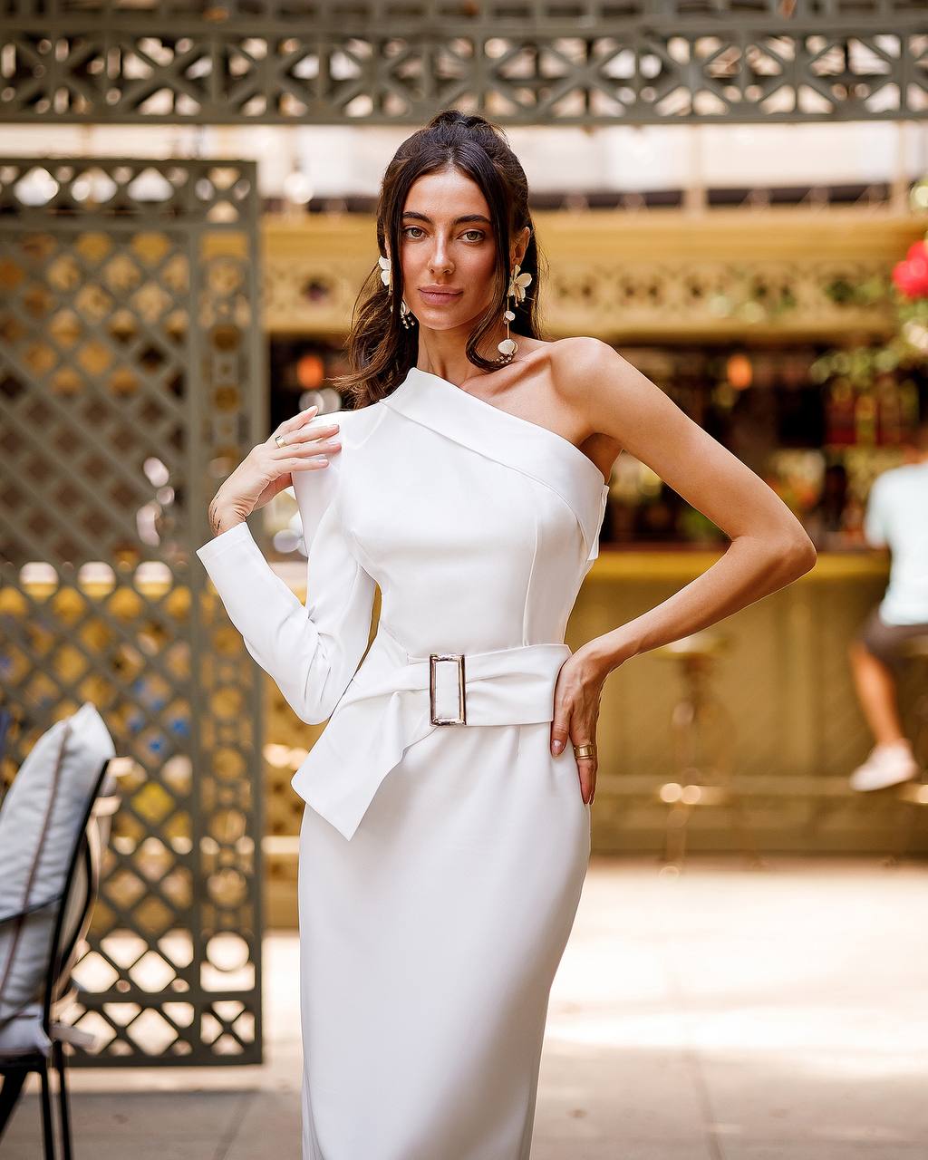 Woman wearing a white one-shoulder dress with a belt in an outdoor setting.
