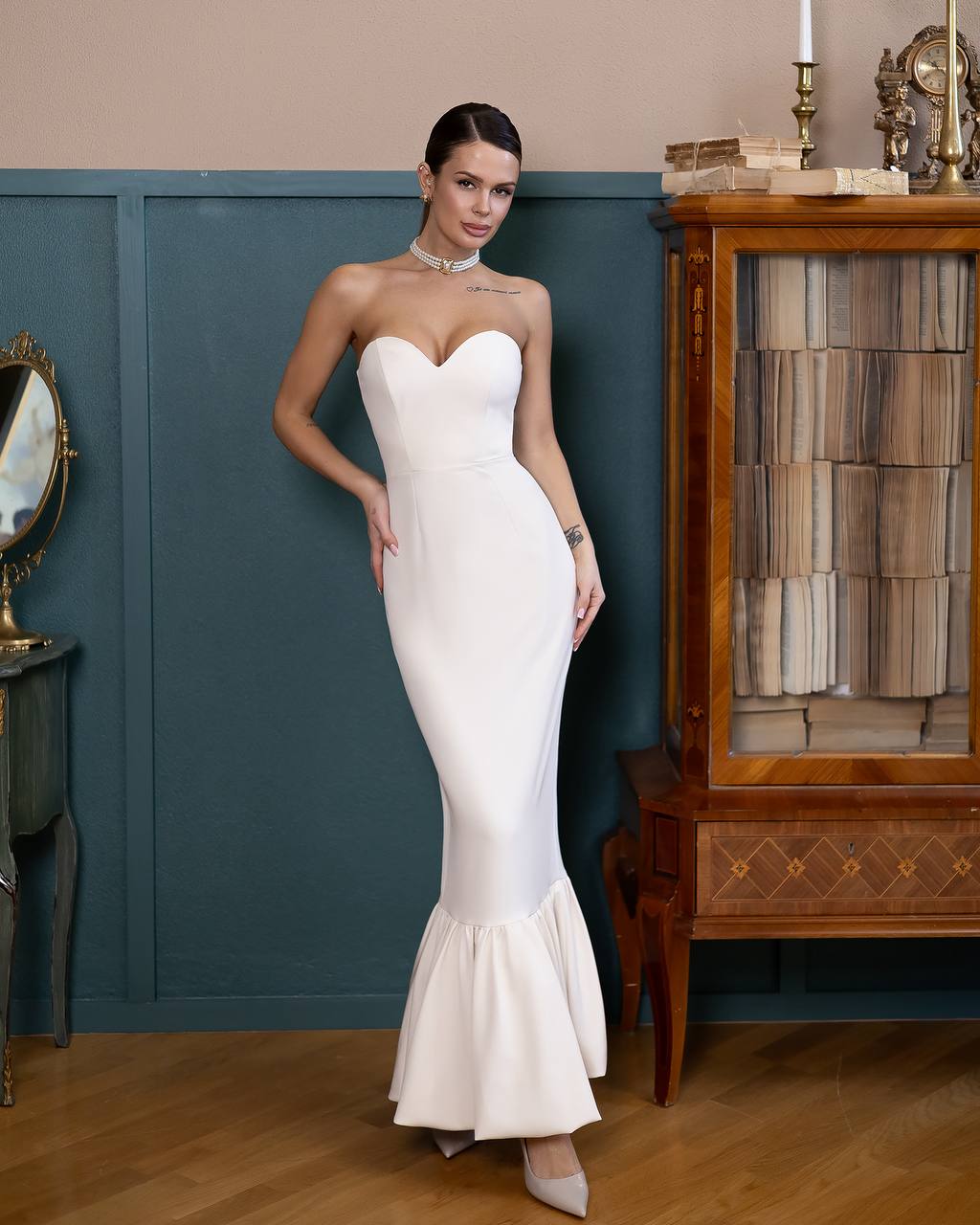 Woman in a white strapless gown standing in an elegant room with wooden furniture and a blue wall.