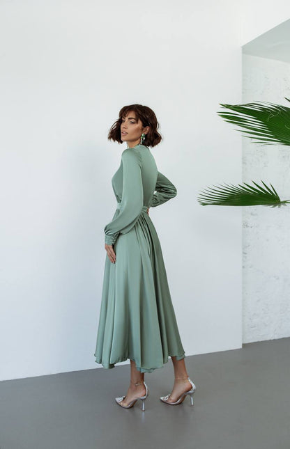 Woman wearing a green dress standing against a white wall with a plant in the corner.