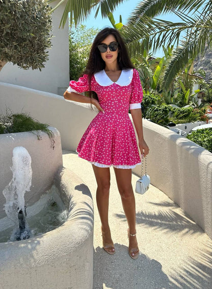 Woman in a pink polka dot dress standing outdoors with palm trees and a fountain in the background