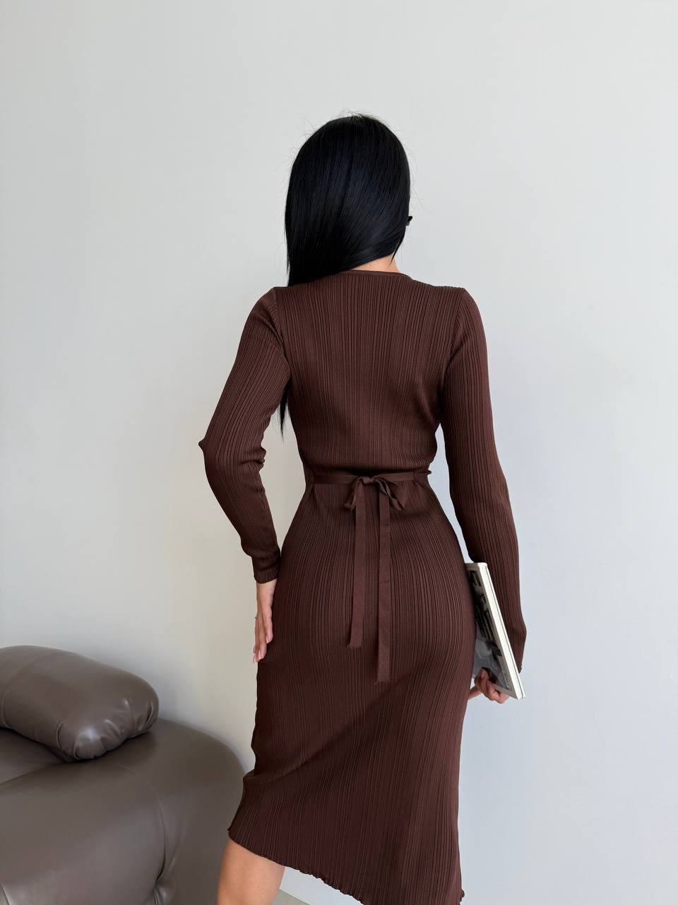 Woman wearing a brown ribbed dress holding a clutch against a plain background