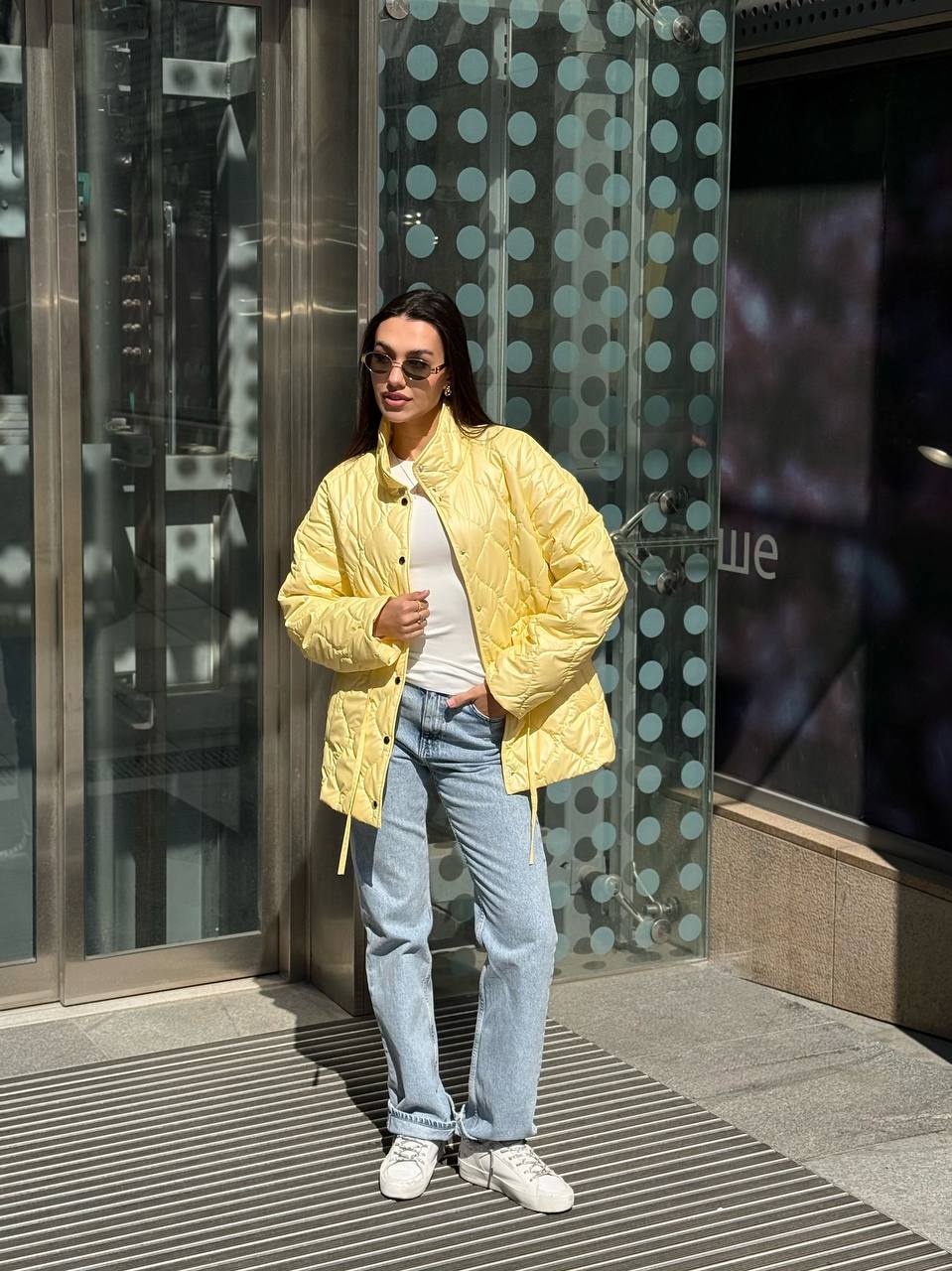 Person wearing a yellow jacket, white shirt, and blue jeans standing in front of a glass door with polka dot pattern.
