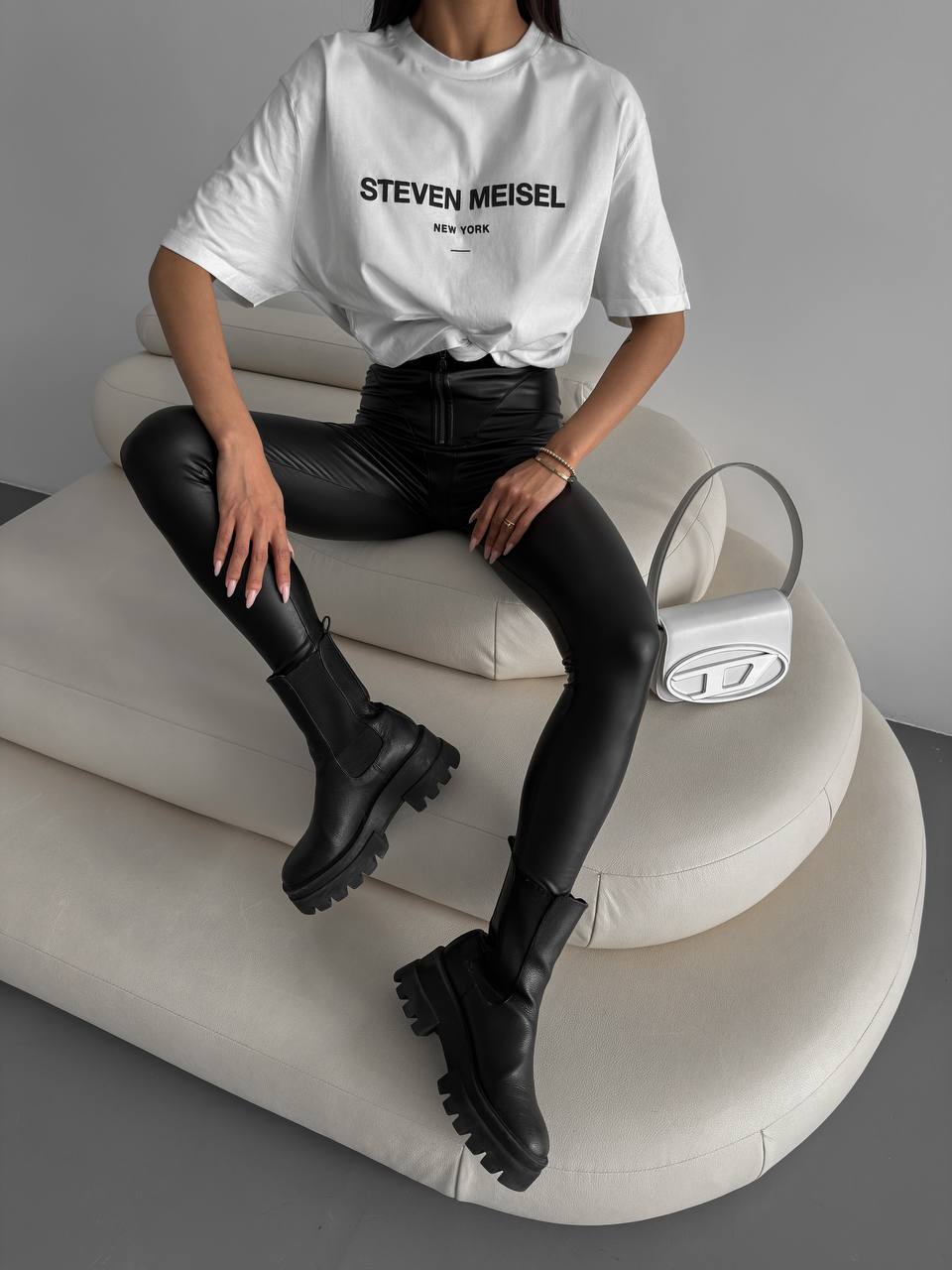 Person wearing a white t-shirt with 'Steven Meisel' branding, sitting on a modern chair.