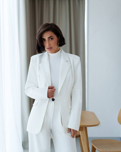 Woman wearing a white suit standing in a room with light-colored curtains and wooden furniture.