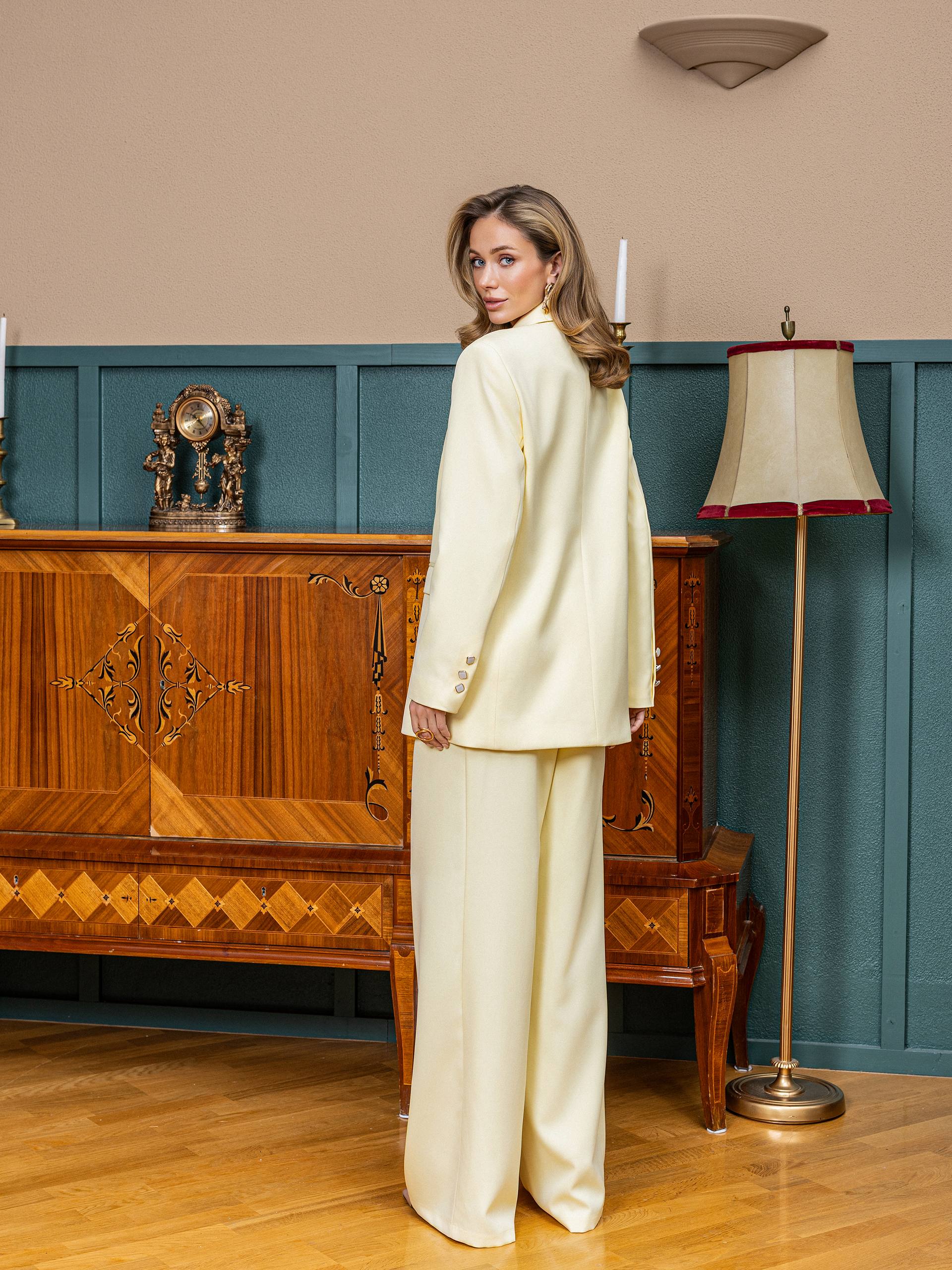 Woman in a cream suit standing in a room with wooden furniture and a lamp.