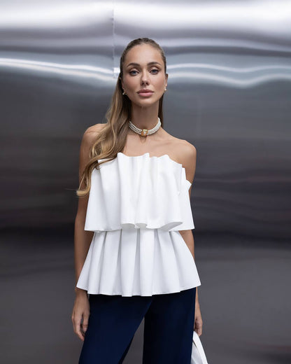 A woman modeling a white off-the-shoulder ruffled peplum top paired with dark navy pants, standing against a metallic silver background.