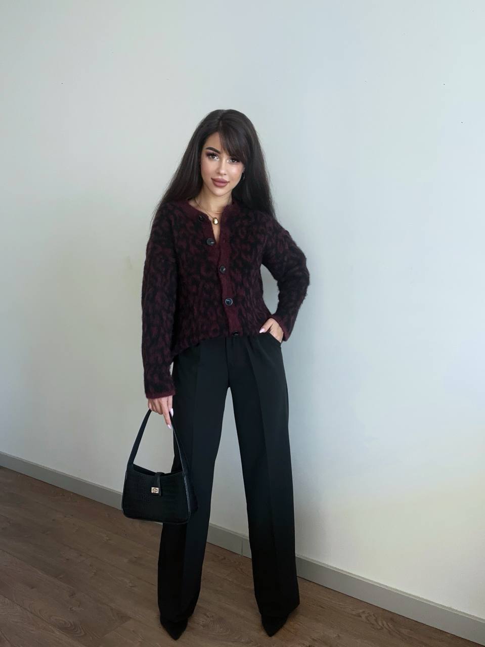 Woman wearing a patterned top and black pants, holding a black handbag against a plain background