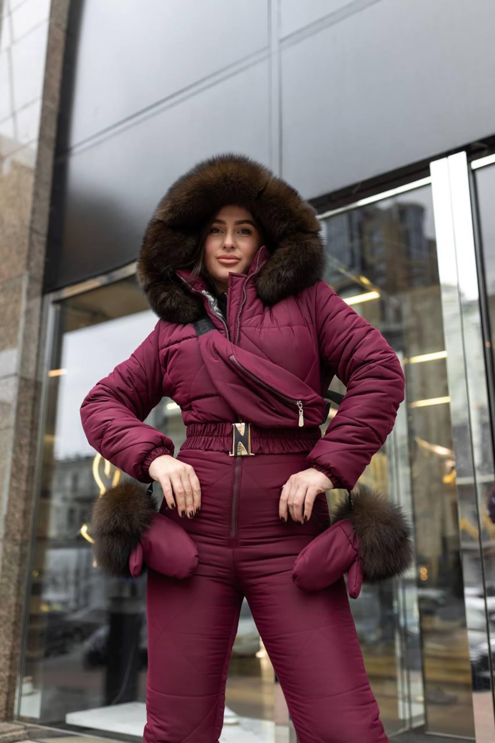 Person wearing a maroon puffer suit with fur trim standing in front of a reflective glass building.