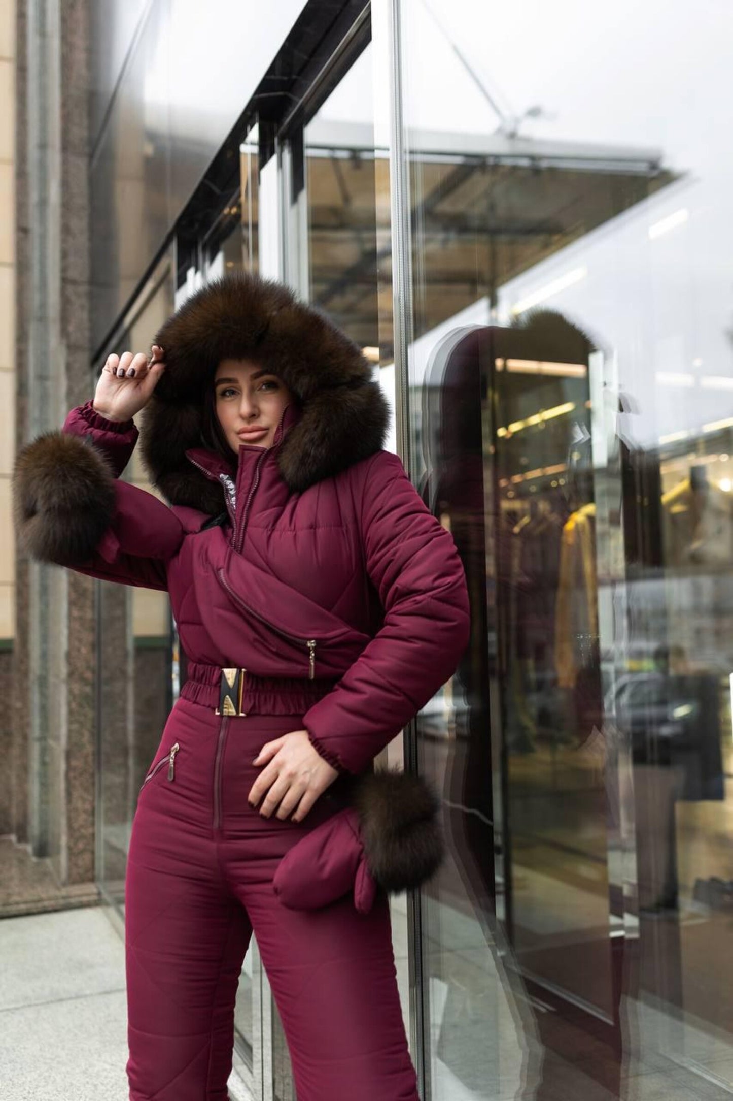 Person wearing a burgundy winter suit with fur trim in front of a reflective glass building.