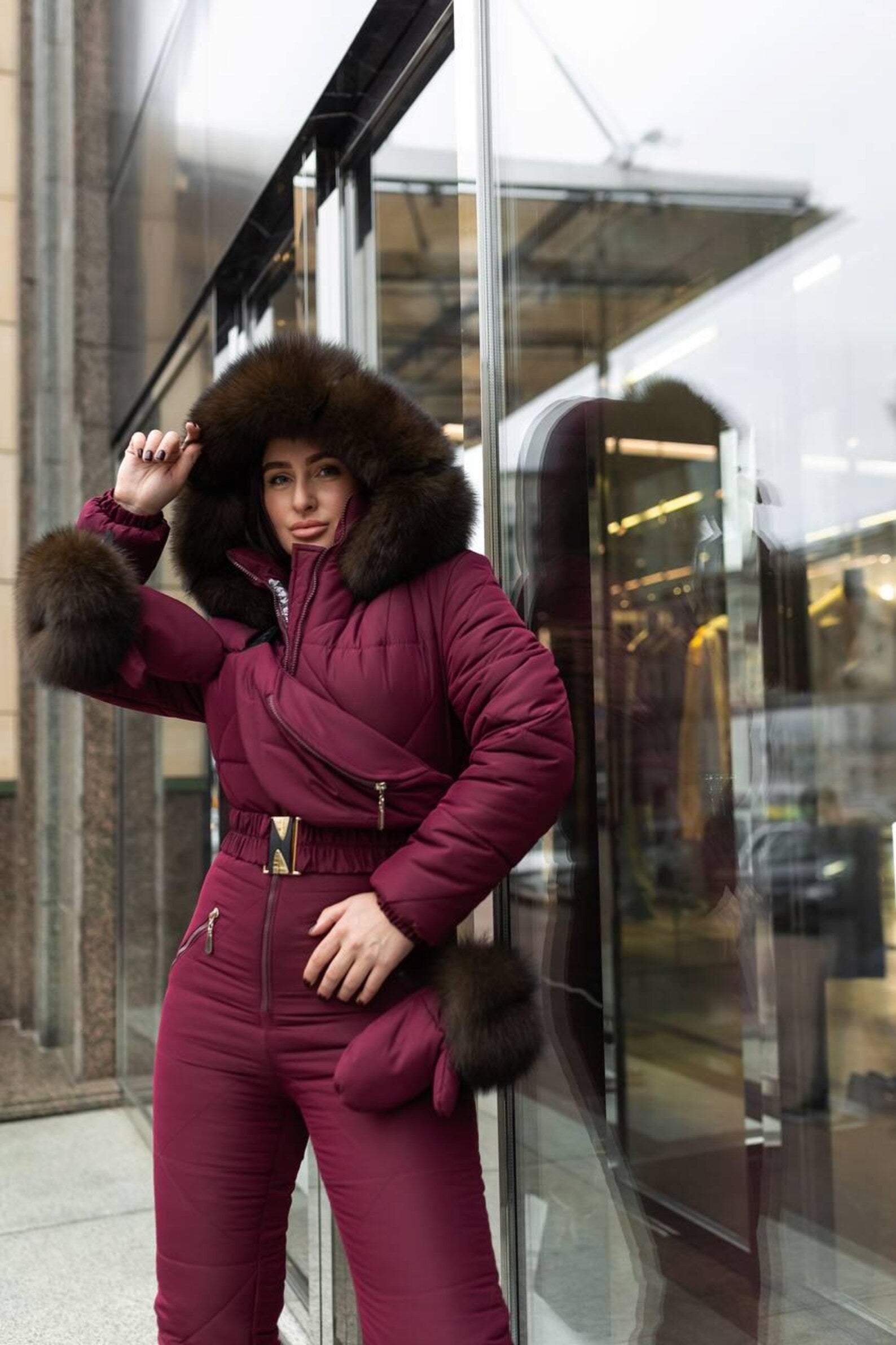 Person wearing a burgundy winter suit with fur trim in front of a reflective glass building.