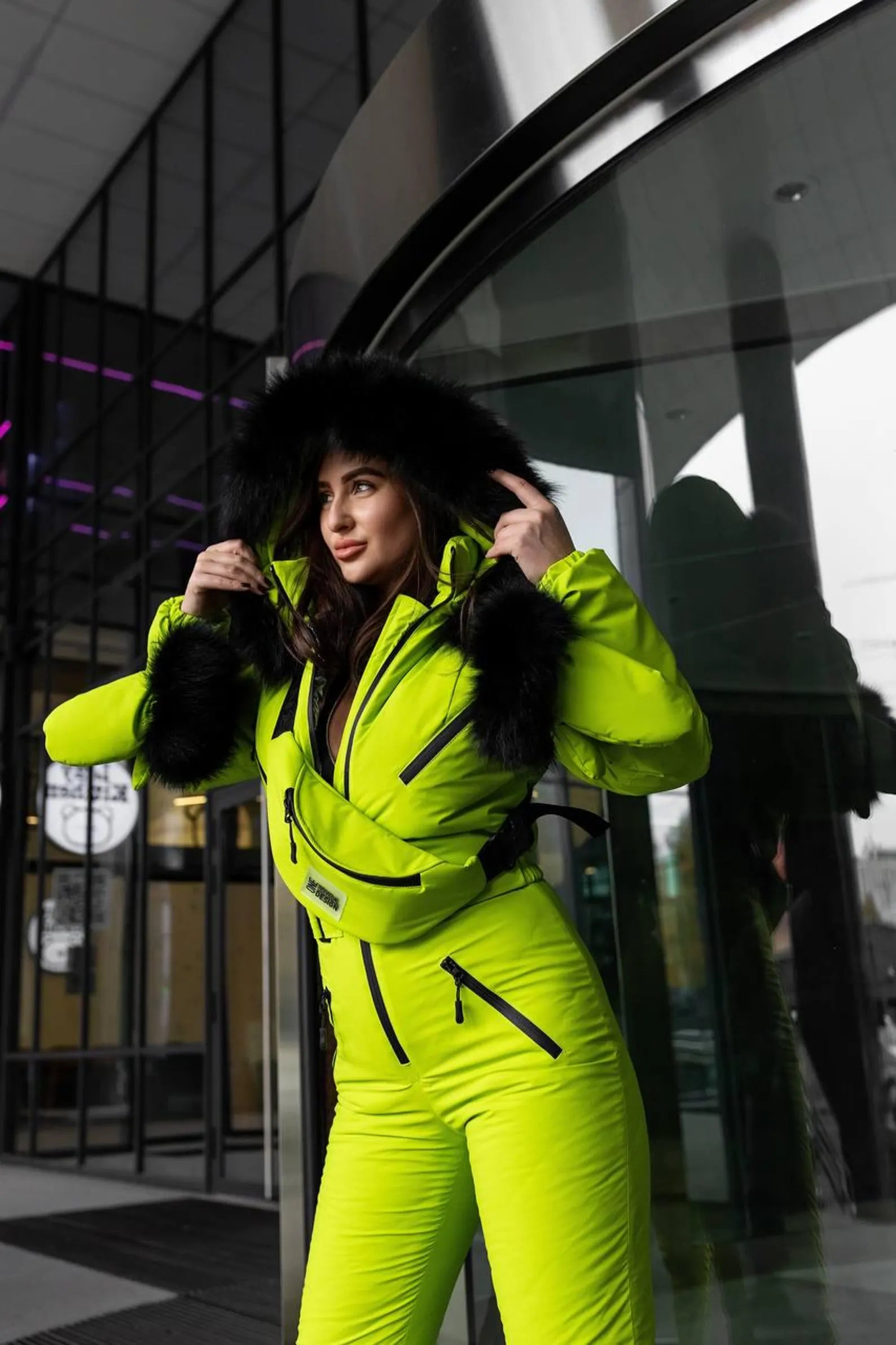 Person wearing a bright yellow snowsuit standing in front of a modern building entrance.