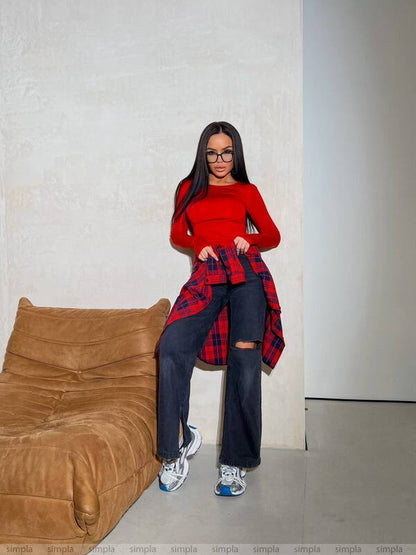 Person wearing a red plaid shirt over a red top with dark jeans, standing in a minimalistic room.