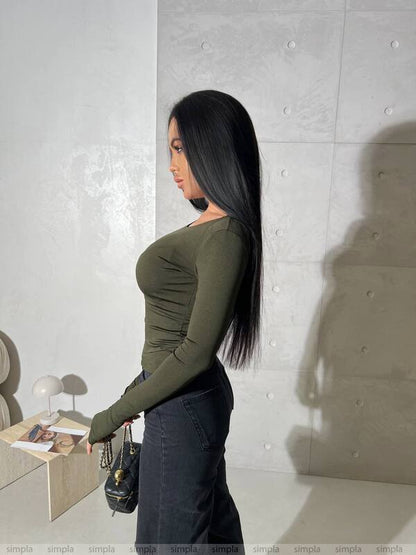 Woman in a green top and dark pants standing against a light gray wall.