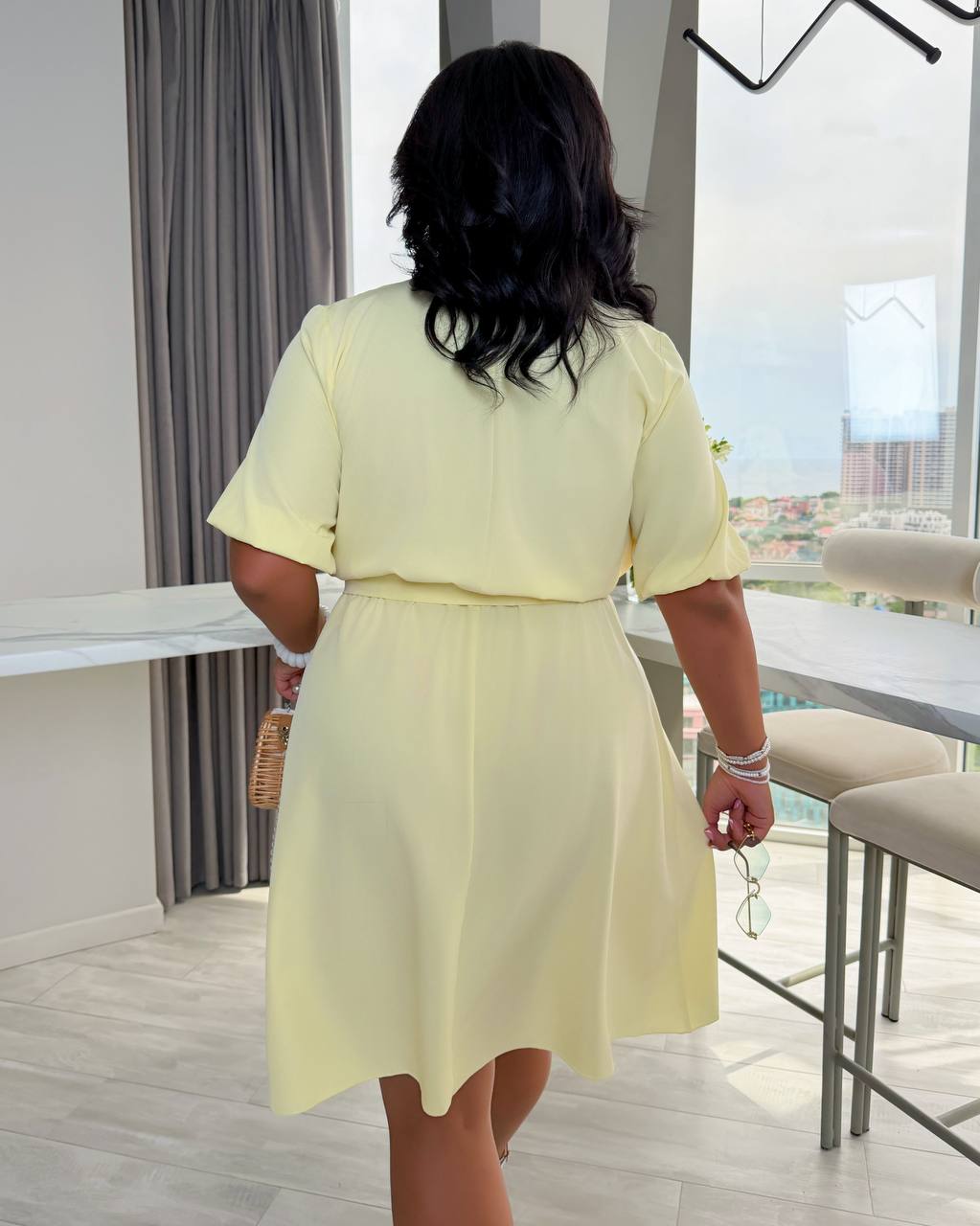 A woman viewed from behind wearing a pale yellow short-sleeve dress in a modern indoor setting with high windows, bar stools, and gray curtains.