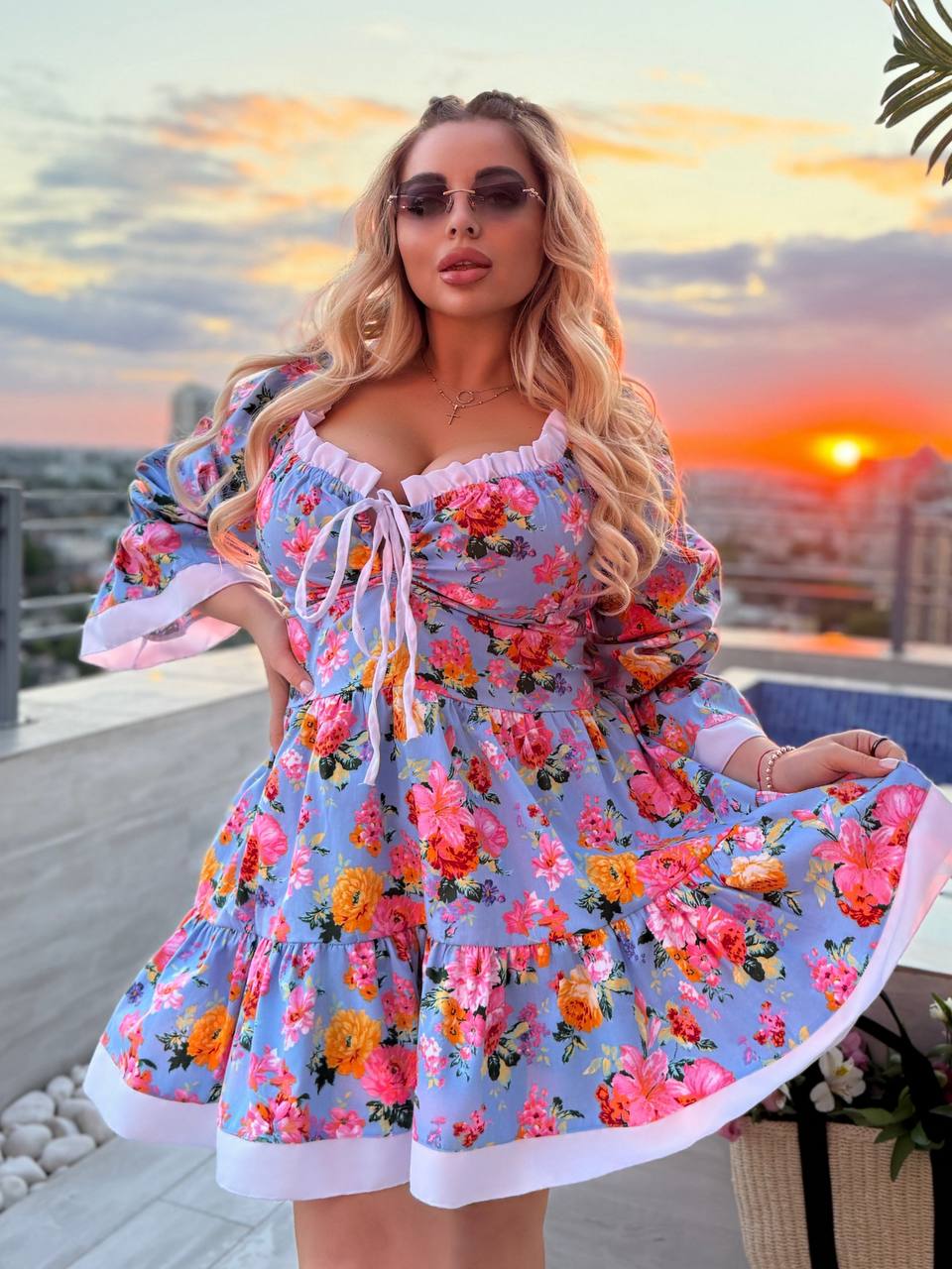 A woman wearing a light blue floral mini dress with ruffled trim and a tie neckline, standing on a rooftop terrace at sunset with a city skyline in the background.