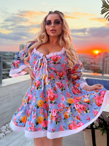 A woman wearing a light blue floral mini dress with ruffled trim and a tie neckline, standing on a rooftop terrace at sunset with a city skyline in the background.