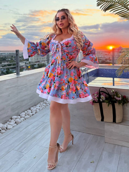 A woman models a short, colorful floral print dress with wide bell sleeves and a white ruffled trim, standing on a rooftop terrace at sunset.