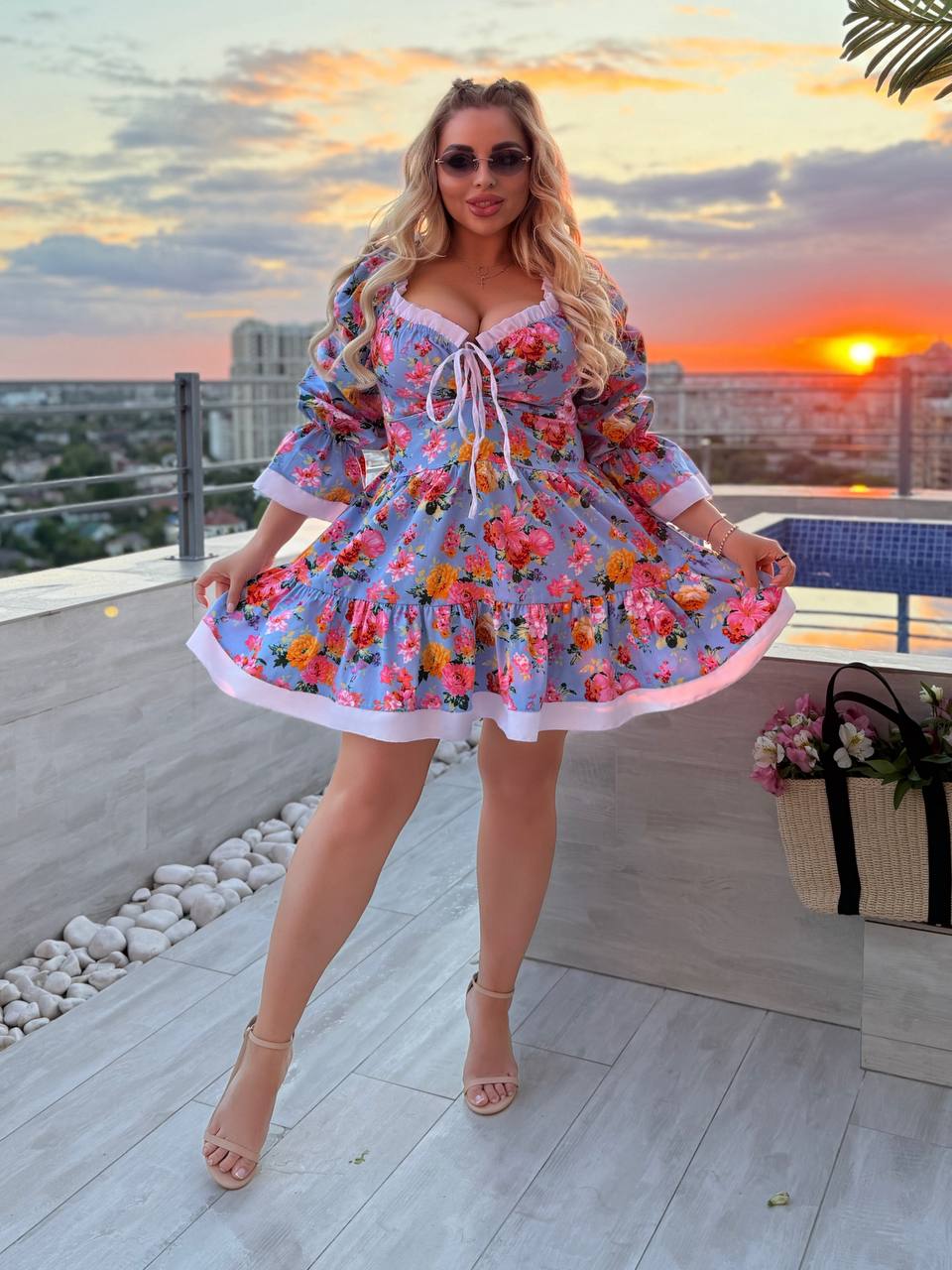 A woman modeling a light blue floral mini dress with a ruffled skirt and tie neckline, standing on a rooftop terrace at sunset.