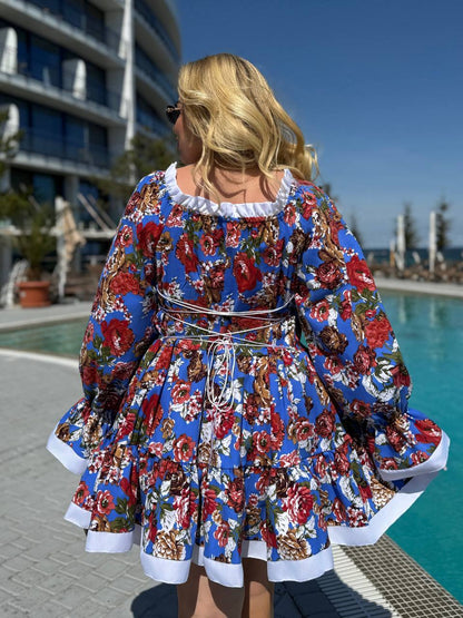 A woman stands with her back to the camera wearing a blue floral dress with white ruffled trim, photographed outdoors beside a swimming pool and modern building.
