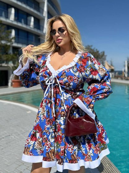 A woman wearing a vibrant blue floral long-sleeve dress with white ruffle trim stands by a swimming pool. She is holding a dark red handbag and wearing sunglasses.