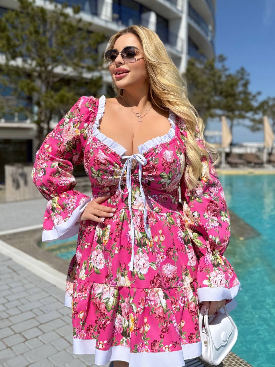 A woman wearing a bright pink floral long-sleeve dress with white ruffle trim and a front tie closure, standing outdoors next to a swimming pool.
