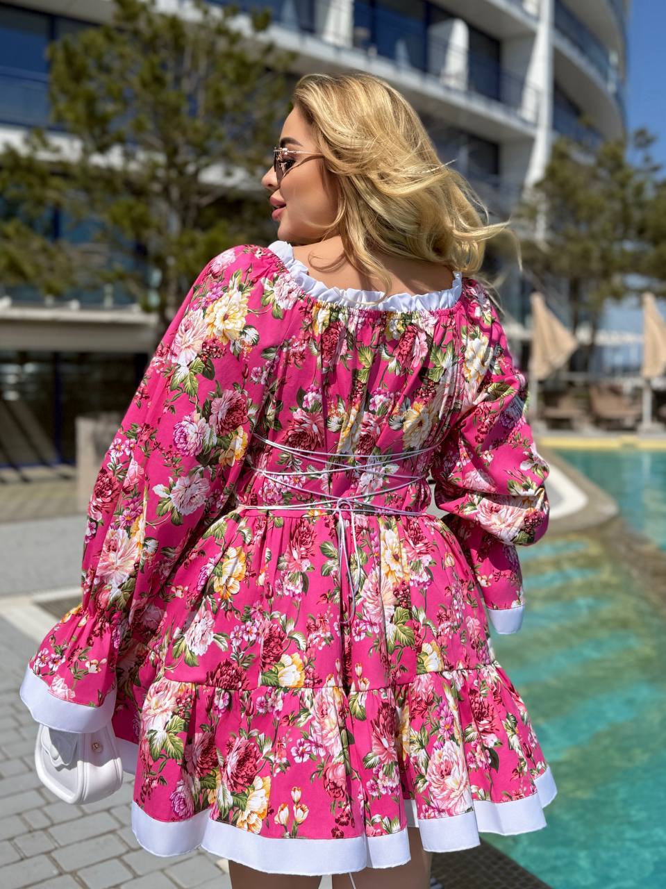 A woman wearing a bright pink floral long-sleeve dress with white ruffle trim, photographed outdoors near a swimming pool and modern building.